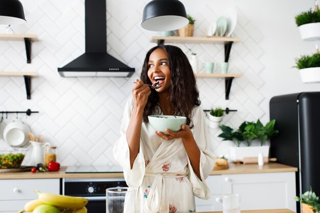 La mujer mulata atractiva sonríe está comiendo frutas cortadas en la moderna cocina blanca vestida con ropa de dormir con el pelo suelto desordenado