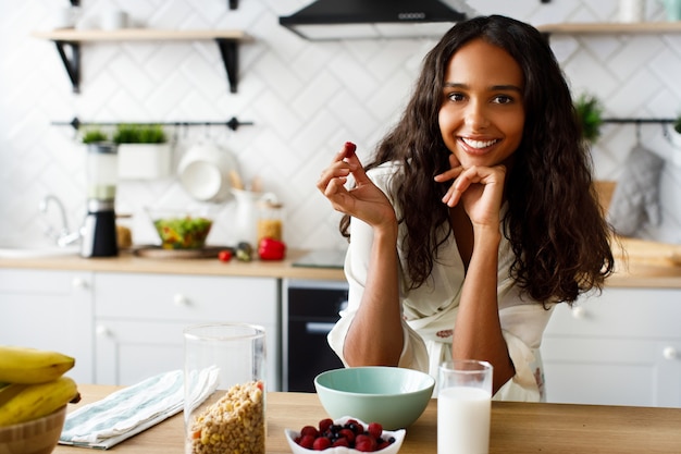 La mujer mulata atractiva sonríe sostiene frambuesa cerca de la mesa con un vaso de leche y crujientes en la moderna cocina blanca vestida con ropa de dormir con el pelo suelto y mirando directamente