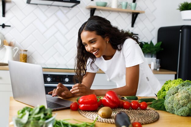 La mujer mulata bonita sonrió está mirando en la pantalla del portátil en la cocina moderna sobre la mesa llena de verduras y frutas, vestida con una camiseta blanca