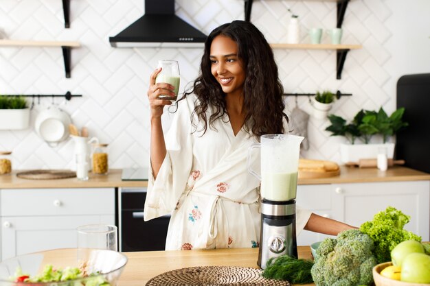 La mujer mulata bonita sonrió sostiene un batido verde cerca de la mesa con verduras frescas en la moderna cocina blanca vestida con ropa de dormir con el pelo suelto y mirando la cristalería