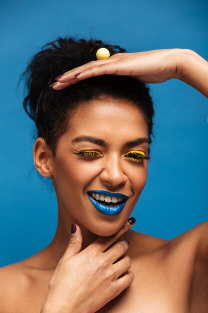 Foto gratuita mujer mulata elegante vertical con maquillaje colorido y cabello rizado en moño posando en cámara con aspecto juguetón aislado, sobre pared azul