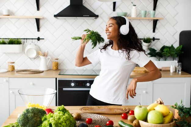 Una mujer mulata guapa con auriculares grandes está sonriendo y fingiendo que está cantando en la vegetación cerca de la mesa con frutas y verduras frescas