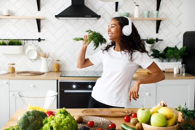 Foto gratuita una mujer mulata guapa con auriculares grandes está sonriendo y fingiendo que está cantando en la vegetación cerca de la mesa con frutas y verduras frescas