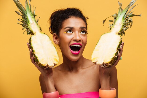 Mujer mulata hambrienta con maquillaje colorido mirando hacia arriba y sosteniendo dos partes de piña fresca apetitosa aislado, sobre pared amarilla