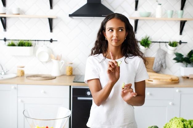 La mujer mulata hermosa consciente está sosteniendo rodajas de limón en la cocina moderna vestida con una camiseta blanca