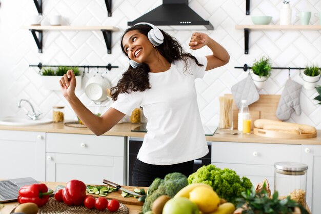 Una mujer mulata sonreída con grandes auriculares inalámbricos baila cerca de una mesa llena de frutas y verduras