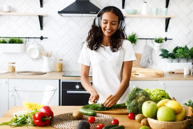 La mujer mulata sonreída con grandes auriculares inalámbricos está cortando vegetación en la cocina moderna junto a la mesa llena de frutas y verduras