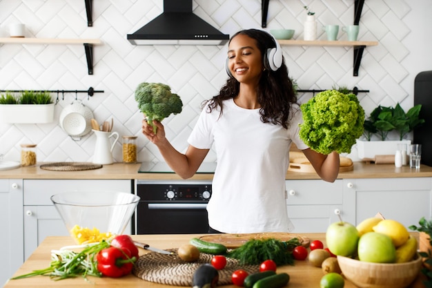 La mujer mulata sonreída con grandes auriculares inalámbricos está sonriendo y sosteniendo ensalada y brócoli en la cocina moderna junto a la mesa llena de frutas y verduras