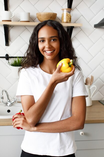 Una mujer mulata sonreída vestida con una camiseta blanca, con cara bonita y cabello suelto sostiene pimientos amarillos en la mano cerca del escritorio de la cocina en la cocina moderna