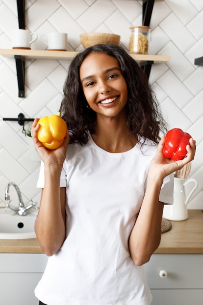 Una mujer mulata sonreída, vestida con una camiseta blanca, con cara bonita y cabello suelto, sostiene pimientos amarillos y rojos en las manos en la cocina