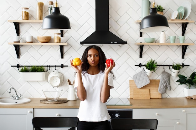 Una mujer mulata vestida con una camiseta blanca, con cara divertida y cabello suelto sostiene pimientos rojos y amarillos en las manos cerca de las mejillas en la cocina moderna