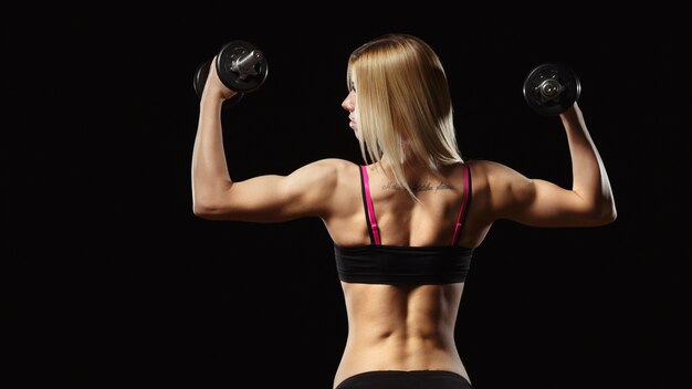 Mujer musculosa de espaldas levantando pesas sobre un fondo negro