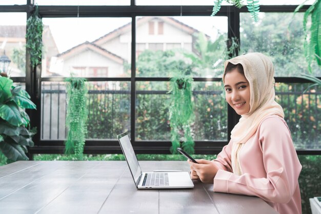 Mujer musulmán asiática sonriente de los jóvenes hermosos que trabaja en el teléfono que se sienta en sala de estar en casa