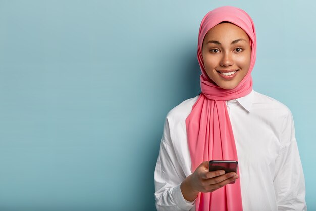 Mujer musulmana complacida usa teléfono celular para socializar, responde en chat en línea, publica algo en redes sociales