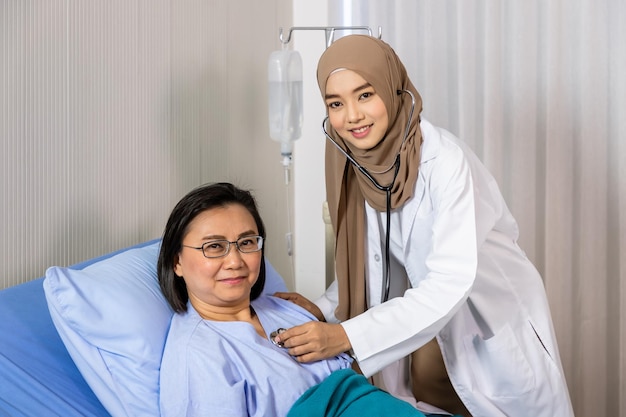 Mujer musulmana doctora en medicina usando estetoscopio para escuchar el latido del corazón del paciente acostado en la cama mirando a la cámara