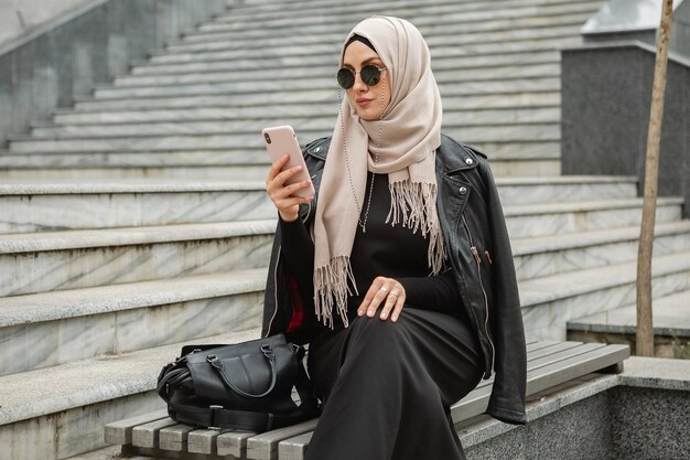 Mujer musulmana con estilo moderno en hijab, chaqueta de cuero y abaya negra caminando en las calles de la ciudad con smartphone