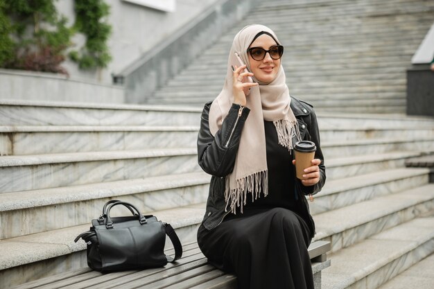Mujer musulmana con estilo moderno en hijab, chaqueta de cuero y abaya negro sentado en la calle de la ciudad hablando por teléfono móvil con gafas de sol