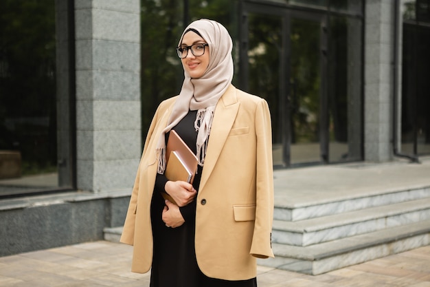 Mujer musulmana con estilo moderno en hijab, chaqueta de estilo empresarial y abaya negra caminando en las calles de la ciudad con laptop