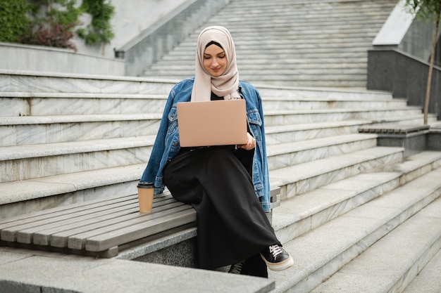 Mujer musulmana con estilo moderno en hijab, chaqueta vaquera y abaya negra sentada en las calles de la ciudad trabajando en la computadora portátil