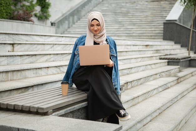 Mujer musulmana con estilo moderno en hijab, chaqueta vaquera y abaya negra sentada en las calles de la ciudad trabajando en la computadora portátil
