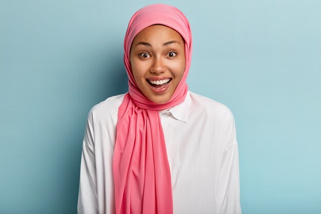 La mujer musulmana llena de alegría emocional se ríe alegremente, tiene una expresión complacida, expresa sentimientos sinceros, usa sombreros rosados, muestra dientes blancos, aislados sobre una pared azul. Concepto de emociones humanas
