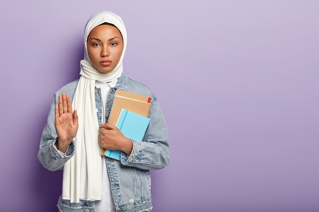 Una mujer musulmana segura y seria sostiene cuadernos de notas, muestra la palma de la mano como señal de rechazo o rechazo, usa una bufanda blanca y un abrigo de mezclilla, pide esperar un minuto, posa sobre una pared púrpura, espacio en blanco