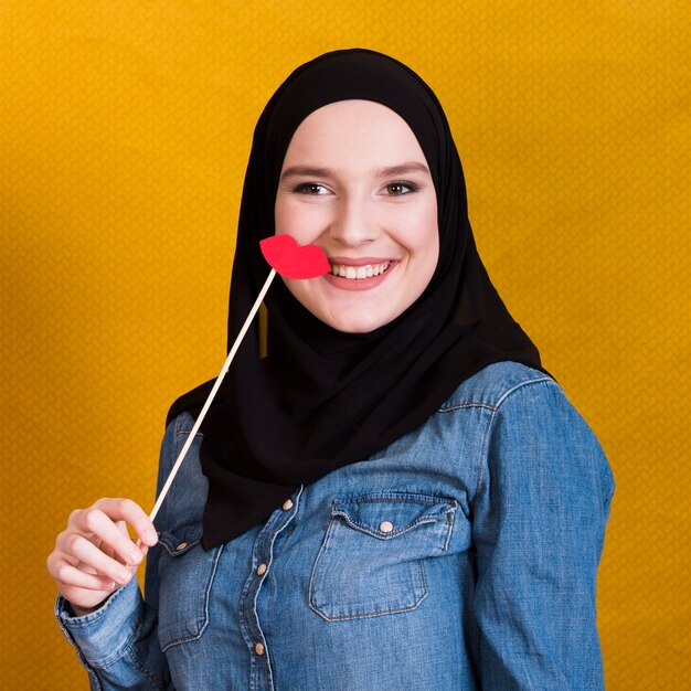 Mujer musulmana sonriente sosteniendo un soporte de papel en forma de labios rojos sobre fondo