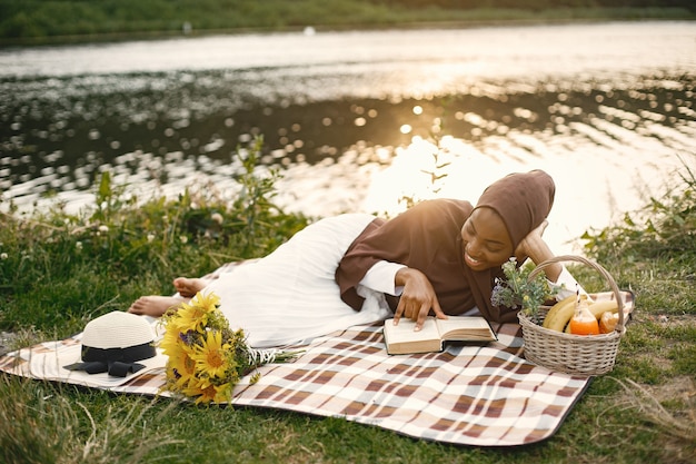 Foto gratuita una mujer musulmana yacía en la manta de picnic a cuadros cerca del río y leía un libro.