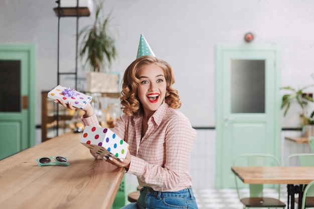 Mujer muy alegre con gorra de cumpleaños sentada en el mostrador del bar con una caja de regalo abierta en las manos y felizmente mirando a un lado en el café