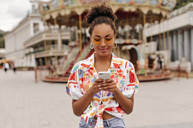 Mujer muy atractiva en pantalones de mezclilla, camisa de colores brillantes y gafas de sol naranjas con teléfono y mensajes afuera