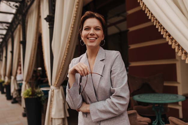 Mujer de muy buen humor mirando a cámara y posando cerca de restaurante. Chica alegre en chaqueta gris de gran tamaño sonríe sinceramente fuera