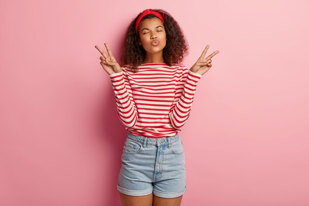 Mujer muy hermosa pacífica con peinado afro, mantiene los labios doblados, hace un gesto de paz con ambas manos, usa un jersey de rayas y pantalones cortos de mezclilla