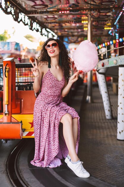 Mujer muy sonriente con cabello oscuro y rizado con gafas de sol y vestido felizmente mirando a un lado mientras se sienta con algodón de azúcar en la mano en un parque de diversiones con atracciones en el fondo