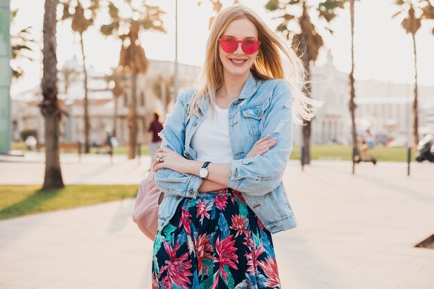 Mujer muy sonriente caminando en las calles de la ciudad con elegante falda estampada y chaqueta vaquera de gran tamaño con gafas de sol rosas, tendencia de estilo veraniego