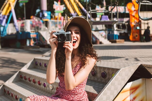 Una mujer muy sonriente con el pelo oscuro y rizado con sombrero sentada y tomando fotos en su pequeña cámara mientras pasa felizmente el tiempo en el parque de diversiones