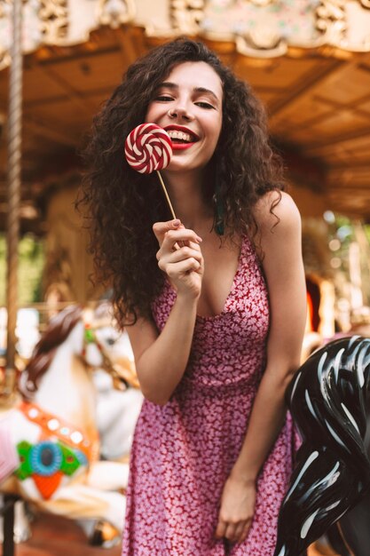 Mujer muy sonriente con el pelo oscuro y rizado vestido de pie con caramelos en la mano y felizmente mirando a la cámara mientras pasa tiempo en el parque de atracciones con carrusel en el fondo