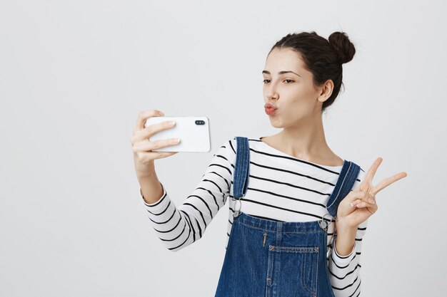 Mujer muy sonriente tomando selfie en smartphone, muestra el signo de la paz