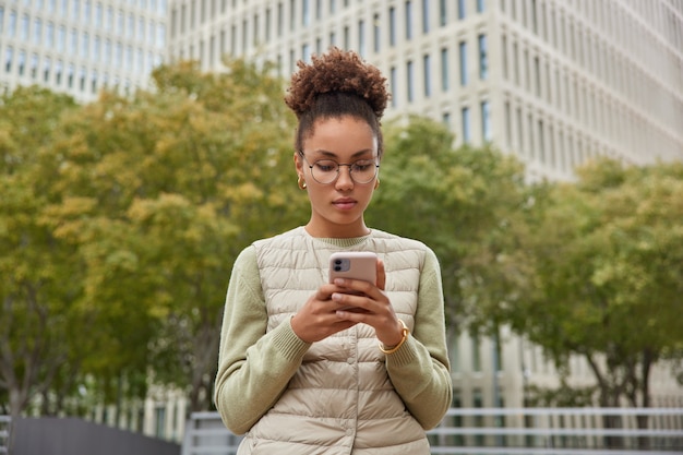 Mujer navega por internet en un teléfono inteligente moderno se apresura a la reunión usa gafas redondas, un jersey y un chaleco posa al aire libre contra un edificio moderno y árboles