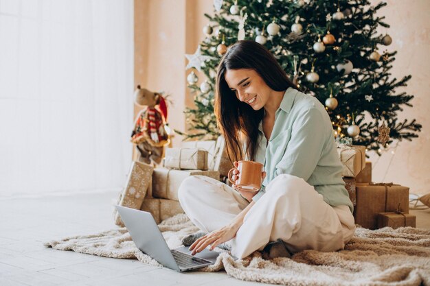 Mujer en Navidad usando laptop y bebiendo té por el árbol de Navidad