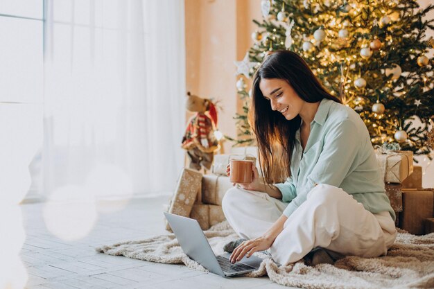 Mujer en Navidad usando laptop y bebiendo té por el árbol de Navidad