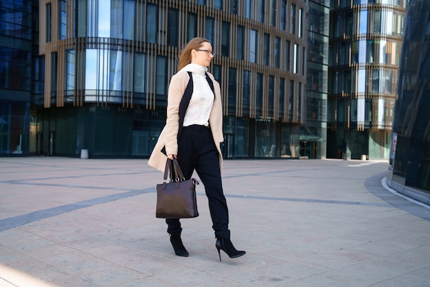 Una mujer de negocios con abrigo y traje, sosteniendo una bolsa en la mano, camina cerca del centro de negocios durante el día. Fotografía horizontal conceptual