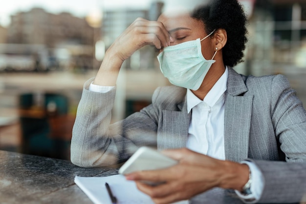 Foto gratuita mujer de negocios afroamericana agotada con mascarilla protectora y sosteniendo la cabeza con dolor la vista es a través del cristal