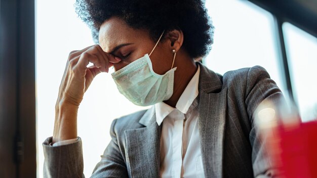 Mujer de negocios afroamericana exhausta con mascarilla facial que tiene dolor de cabeza mientras está sentada en un café