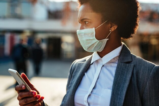 Mujer de negocios afroamericana con máscara protectora en la cara mientras usa un teléfono inteligente en la ciudad