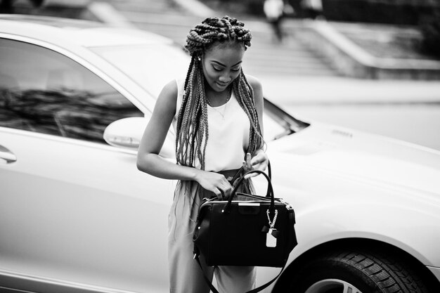 Mujer de negocios afroamericana de moda en pantalones rosas y rastas rojas posadas al aire libre con una bolsa contra su auto de lujo