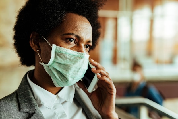 Mujer de negocios afroamericana pensativa que usa mascarilla mientras habla por teléfono en la estación de tren pública durante la epidemia de virus