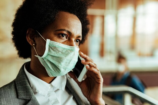 Mujer de negocios afroamericana pensativa que usa mascarilla mientras habla por teléfono en la estación de tren pública durante la epidemia de virus