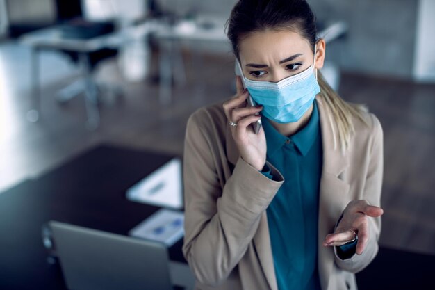 Mujer de negocios angustiada con mascarilla hablando por teléfono en la oficina