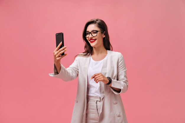 Mujer de negocios en anteojos y traje toma selfie sobre fondo rosa. Chica encantadora alegre con cabello largo oscuro con lápiz labial rojo hace foto.