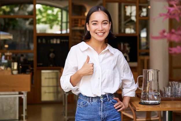 Foto gratuita mujer de negocios asiática confiada que muestra los pulgares hacia arriba de pie cerca de la entrada de su café o restaurante rec